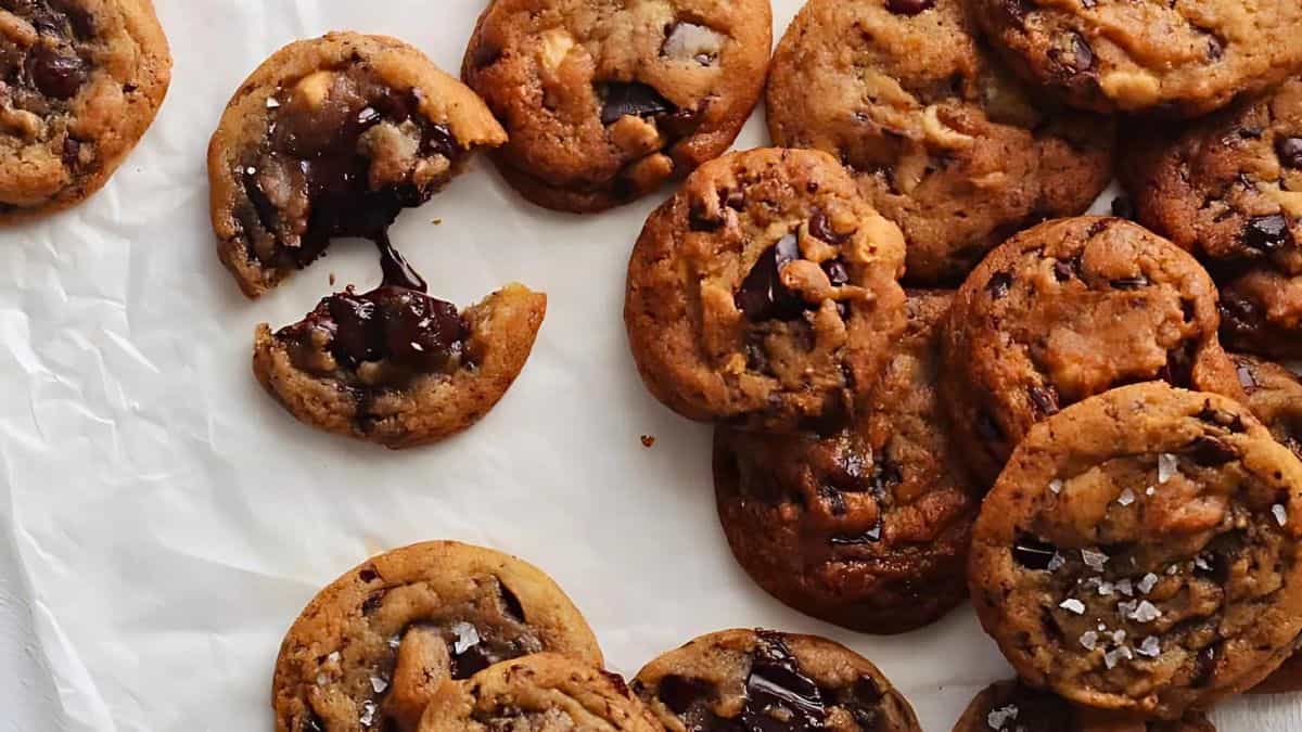 A batch of chocolate chip cookies, some topped with sea salt. One cookie is broken in half, revealing melted chocolate inside. They are arranged on white parchment paper.