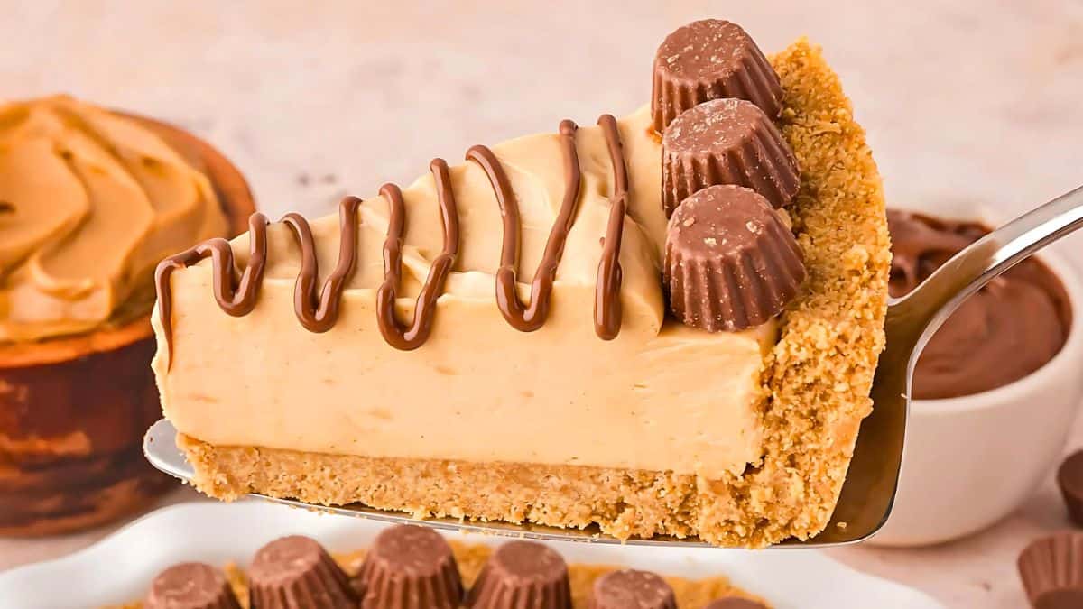 A slice of peanut butter pie with a graham cracker crust is topped with mini chocolate peanut butter cups and drizzled with chocolate. The pie serves as a background with additional bowls of peanut butter and chocolate.