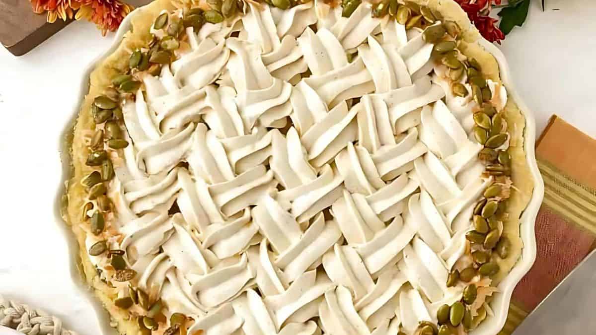 A pie with a woven lattice crust is topped with cream and garnished with pumpkin seeds around the edges. It rests on a table with autumn flowers and a knit cloth nearby.