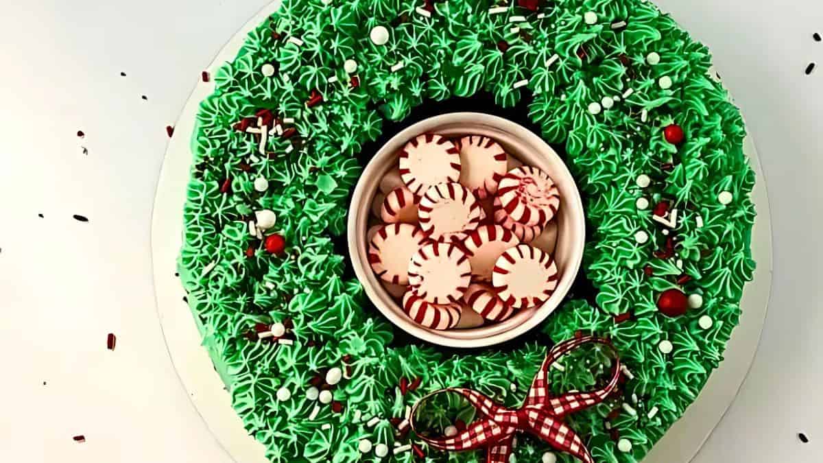 A festive cake decorated like a Christmas wreath with green frosting, red and white beads, and a checked ribbon. The center has peppermint candies displayed in a small dish.