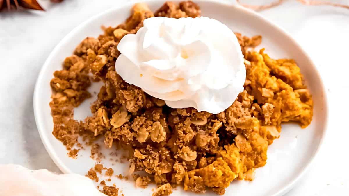 A plate of pumpkin crumble topped with a dollop of whipped cream. The crumble has a golden-brown, crumbly topping over a layer of pumpkin filling. The plate is set on a white surface.