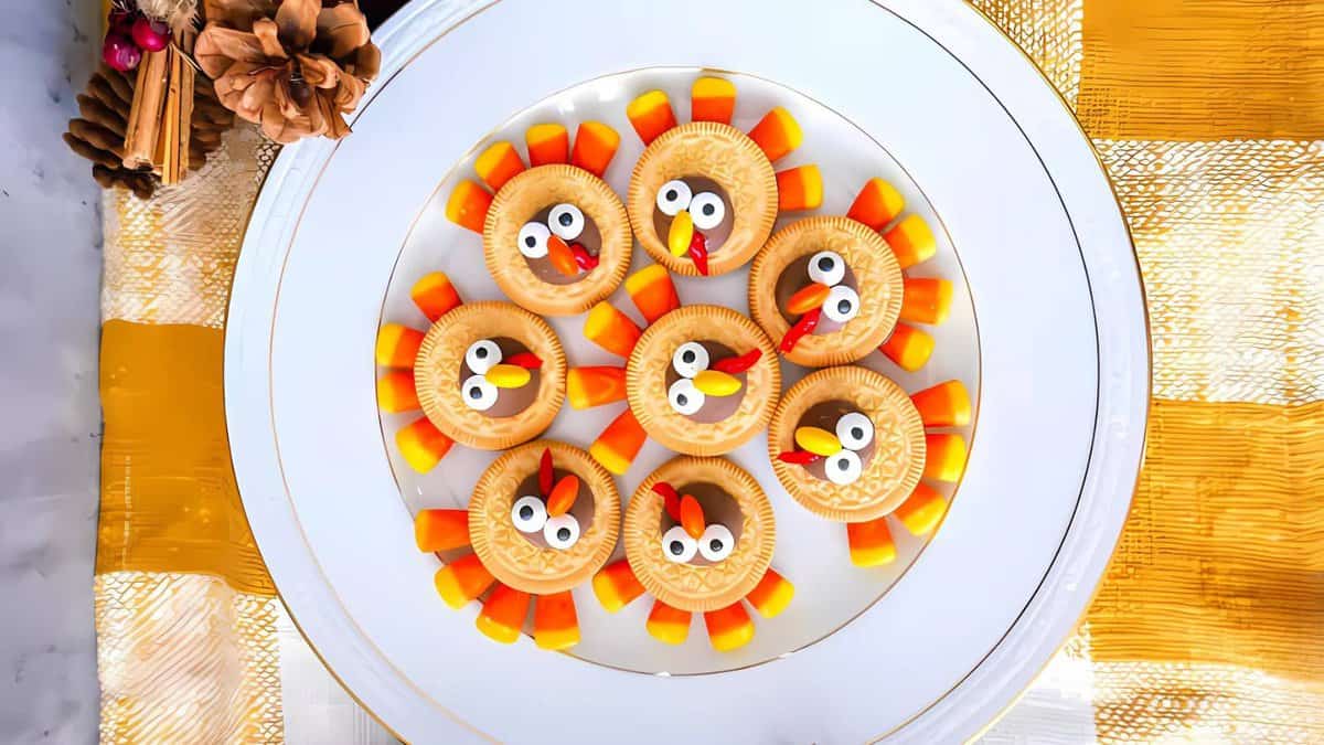 A plate of Thanksgiving cookies, cleverly designed to resemble turkeys with candy eyes, candy corn for feathers, and red candy beaks, all neatly arranged in a circular pattern. The background accentuates the festive feel with a decorative tablecloth and cinnamon sticks.