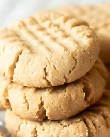 A close-up of several peanut butter cookies stacked on a light surface. The cookies have a crisscross pattern on top, suggesting a classic homemade style. They appear crumbly and golden brown.