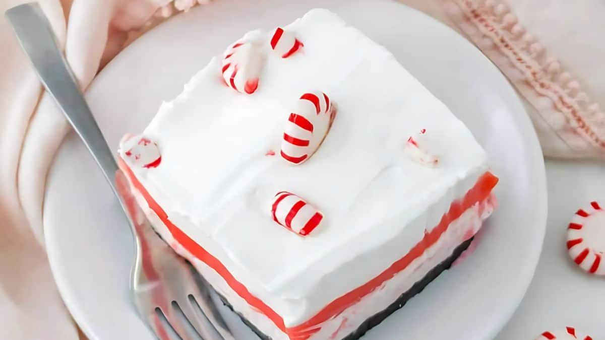 A slice of layered dessert with a white whipped topping on a plate. It's garnished with red and white peppermint candies. A fork rests on the side of the plate. The background features a soft, pink fabric.