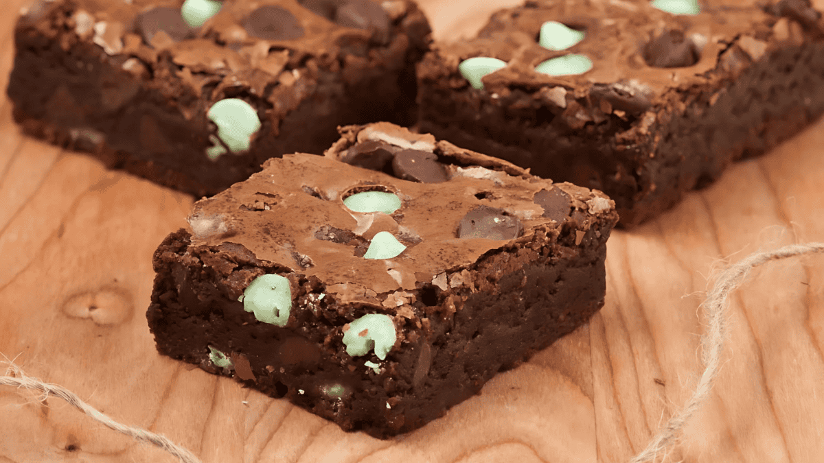 Four chocolate brownies with mint chips are arranged on a wooden surface. The brownies have a rich, fudgy texture and are dotted with green mint chips and chocolate chunks, creating a visually appealing contrast.
