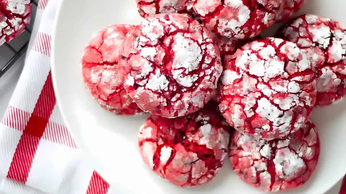 A white plate filled with red crinkle cookies dusted with powdered sugar. A red and white checkered cloth is partially visible beside the plate. The cookies have a cracked surface revealing a vibrant red color.