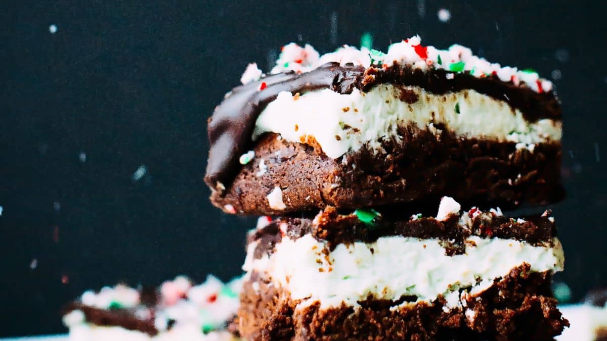 A close-up of two stacked, chocolate brownies topped with a layer of white frosting and sprinkled with crushed peppermint candy. The background is dark, highlighting the vibrant colors of the dessert.