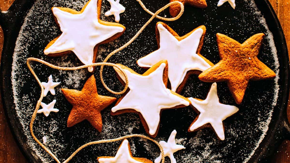 A round black tray is filled with star-shaped cookies, some with white icing and some plain brown. The cookies are creatively arranged with a string of smaller stars. Dusting of powdered sugar adds a festive touch.