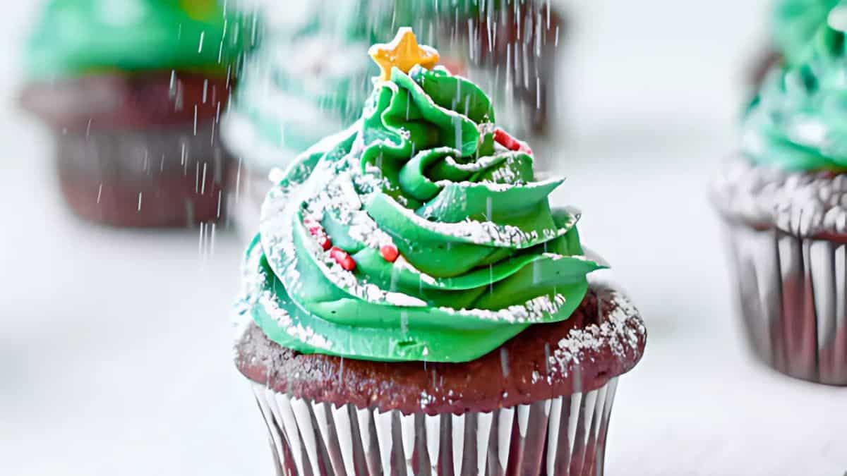 A chocolate cupcake topped with green frosting shaped like a Christmas tree, decorated with a yellow star and red sprinkles. Powdered sugar is being sprinkled on top, resembling snow. Other cupcakes are in the blurred background.