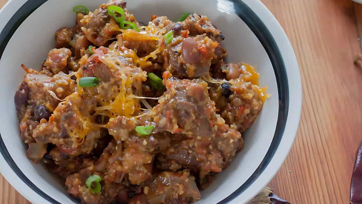 A bowl of chili featuring chunks of meat, beans, and a tomato-based sauce is garnished with melted cheese and chopped green onions, placed on a wooden table.