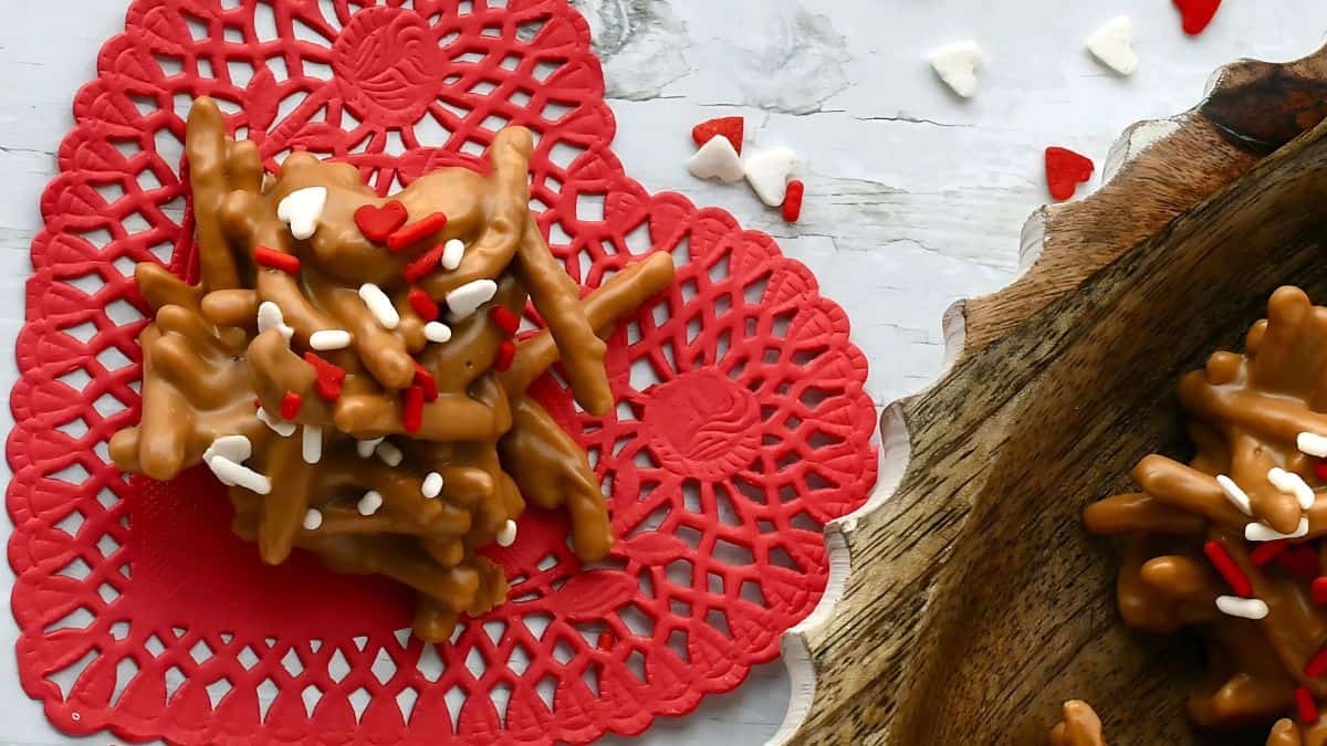 A cluster of caramel-coated treats topped with red and white heart-shaped sprinkles sits on a red heart doily. Nearby, a wooden tray contains more treats. White and red heart sprinkles are scattered on a light surface.