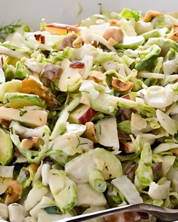 A bowl of fresh coleslaw with sliced apples, cashews, and raisins, mixed with a creamy dressing. A silver spoon rests in the bowl, ready to serve.