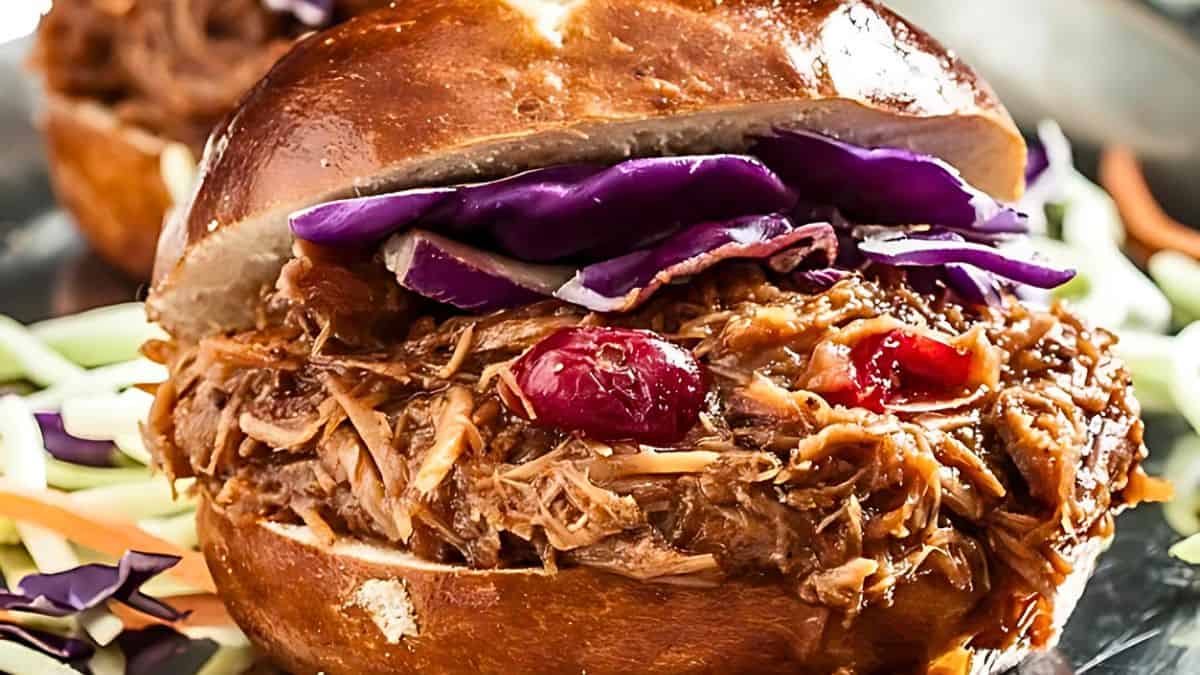 A savory pulled pork sandwich on a shiny bun, topped with vibrant slices of red cabbage. Background features colorful shredded vegetables, adding to the appetizing presentation.