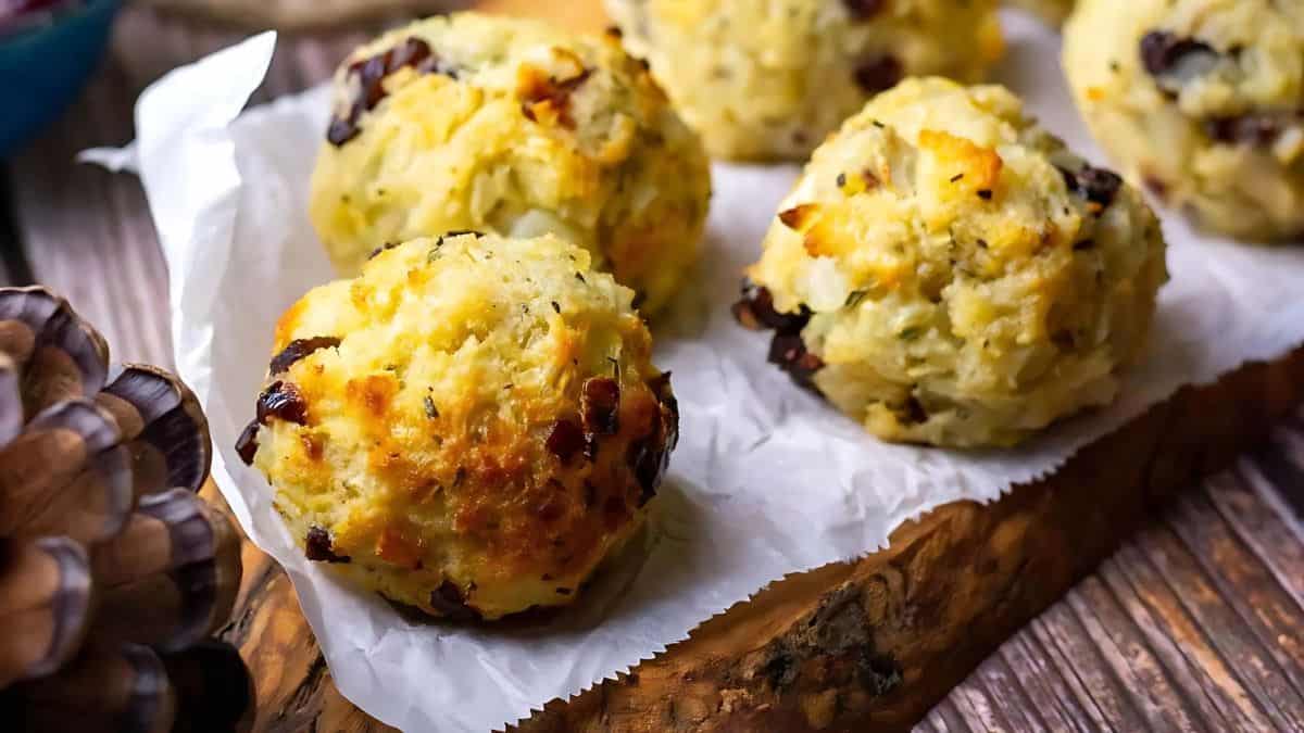 Four golden-brown baked potato balls with cheese and herbs sit on a piece of parchment paper on a rustic wooden board. They are dotted with crispy bits and look freshly baked.