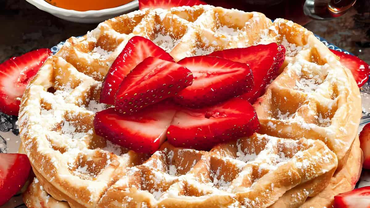 A close-up of a waffle topped with sliced strawberries and a dusting of powdered sugar. Additional strawberry slices are scattered around the waffle. In the background, there are small bowls containing syrup and honey.