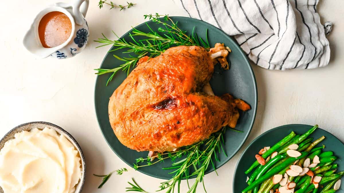A roasted turkey garnished with rosemary sits on a plate. Surrounding it are dishes of green beans topped with almonds, creamy mashed potatoes, and a gravy boat. A striped kitchen towel is nearby.