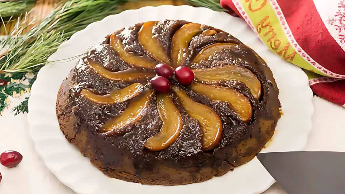 A delicious upside-down cake with caramelized apple slices arranged in a circular pattern on top. Three red cherries adorn the center. The cake is on a white plate, accompanied by a festive red and white cloth and green sprigs.