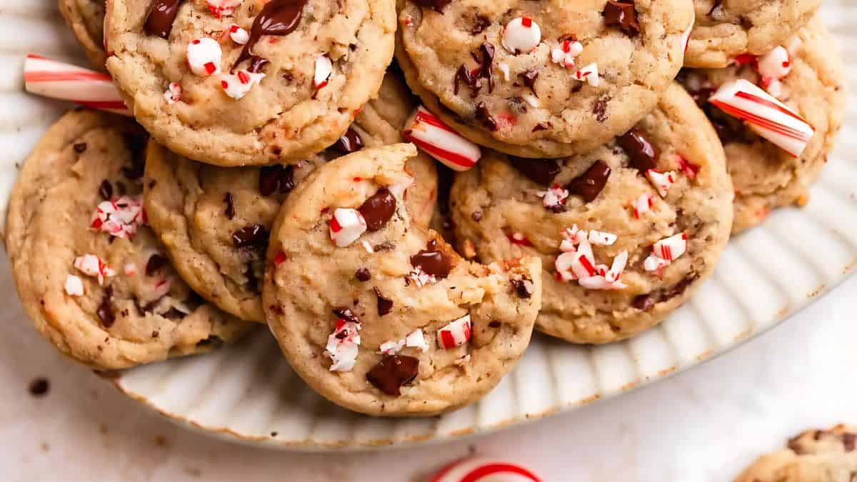 A plate of chocolate chip cookies topped with crushed candy canes. The cookies appear soft and chewy, with chocolate chunks and minty pieces scattered on top. Several whole candy canes are placed decoratively around the plate.