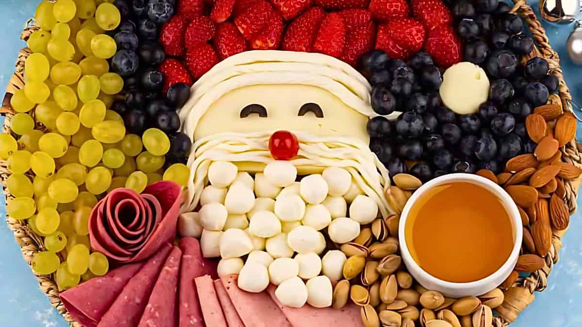A festive platter shaped like Santa Claus, featuring mozzarella balls as a beard, blueberries as a hat, strawberries as a hat trim, and various meats and cheeses arranged creatively. A small cup of honey is placed on the right side.