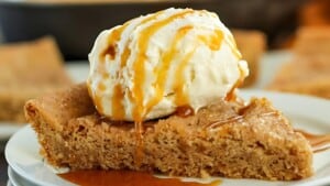 A slice of brown sugar cake topped with a scoop of vanilla ice cream, drizzled with caramel sauce, sits on a white plate. The cake is crumbly and moist, and the ice cream is slowly melting over the warm dessert.