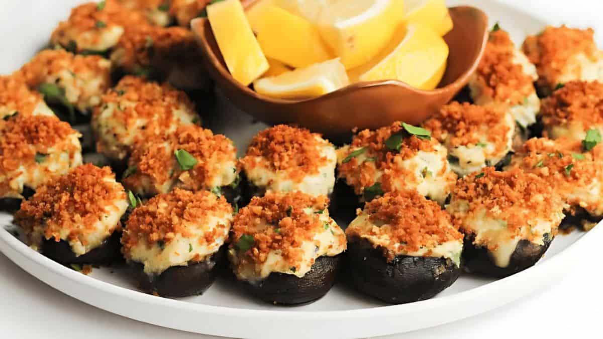 A plate of stuffed mushrooms topped with crispy breadcrumb and herb mixture. In the center, a small dish contains lemon wedges. The presentation is neat and appetizing.
