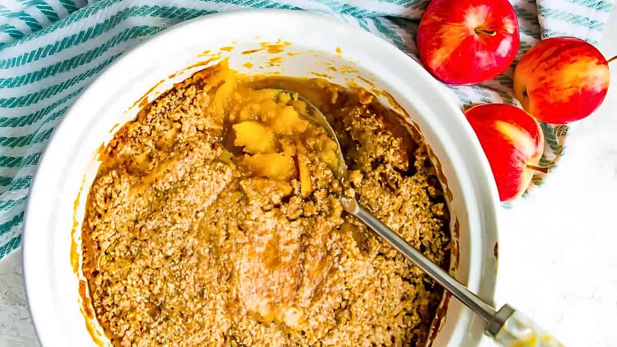 A white baking dish with a spoon scooping apple crumble, featuring a golden-brown crumbly topping and soft apple filling. Three red apples and a striped cloth are beside the dish on a white surface.