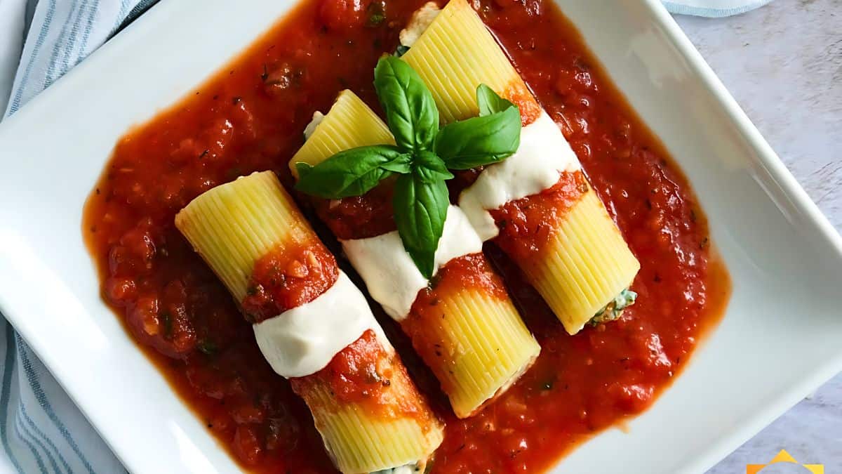 Three manicotti pasta tubes filled with ricotta cheese and spinach on a white square plate, topped with tomato sauce and mozzarella, garnished with fresh basil leaves. The dish is served on a light-colored table with a striped napkin.