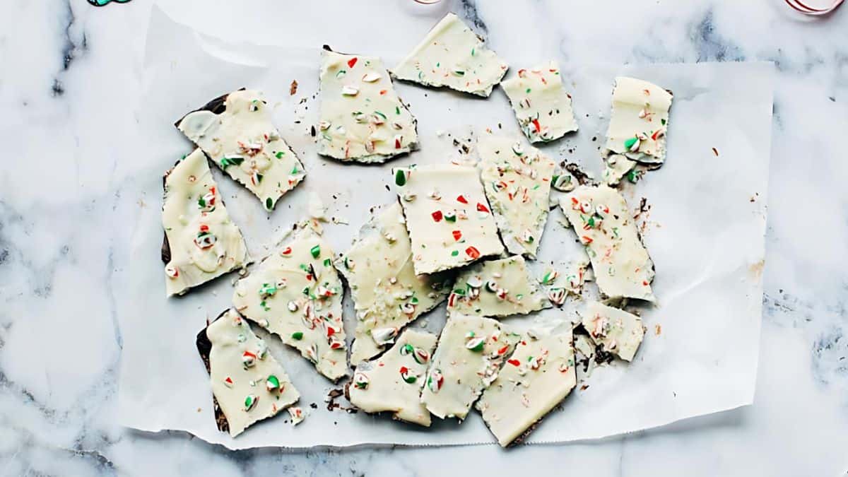 Pieces of peppermint bark with crushed candy canes on top, arranged on parchment paper against a marble background. The bark has a layer of white chocolate with peppermint pieces.