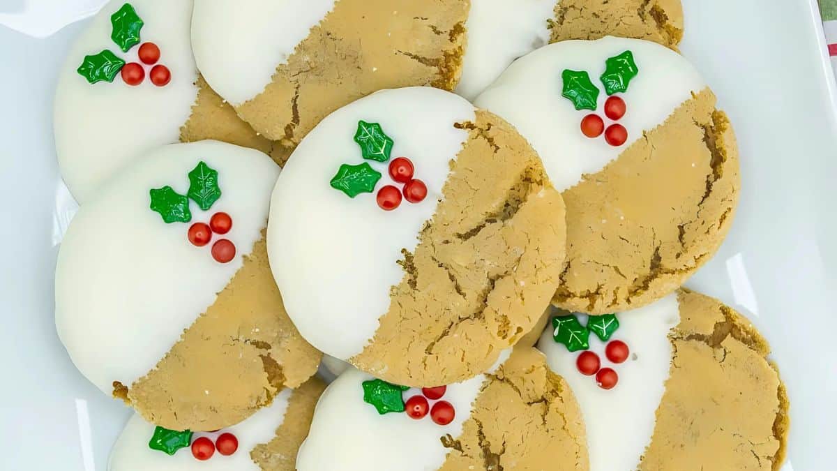 A plate of cookies dipped halfway in white chocolate, adorned with tiny red candies and green leaf decorations, resembling holly. The cookies are arranged in a festive presentation.