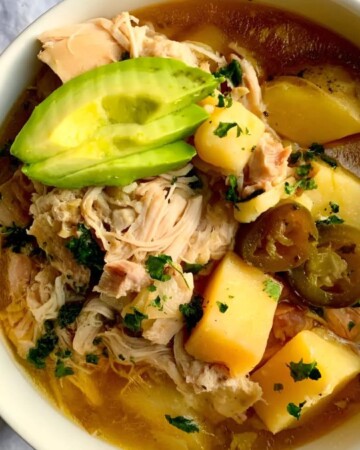 A bowl of warm chicken soup served with chunks of potatoes, sliced avocado, and jalapeño slices, garnished with fresh herbs. It's placed on a white cloth on a marble surface.