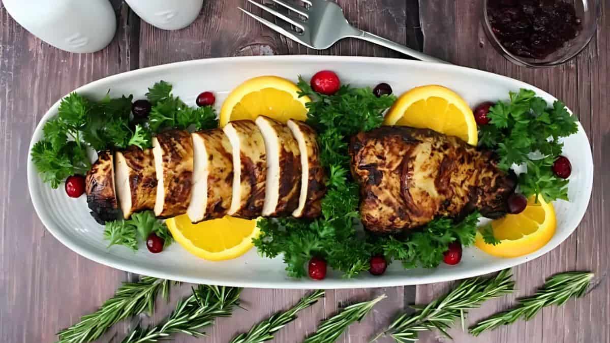 A beautifully plated dish featuring grilled turkey breast slices on a white oval platter, garnished with fresh parsley, orange slices, and cranberries. Nearby are rosemary sprigs and a fork on a rustic wooden table.