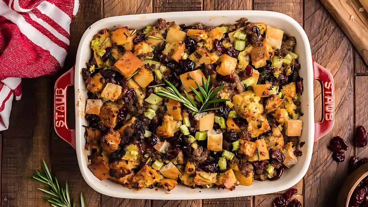 A casserole dish filled with a holiday stuffing mix, featuring chunks of bread, butternut squash, cranberries, celery, and herbs, garnished with a rosemary sprig. The dish rests on a wooden table beside a red-striped cloth and a sprig of rosemary.