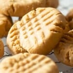 Close-up of several peanut butter cookies with fork marks on top, resting on a cooling rack. The cookies have a golden-brown color and a crumbly texture.