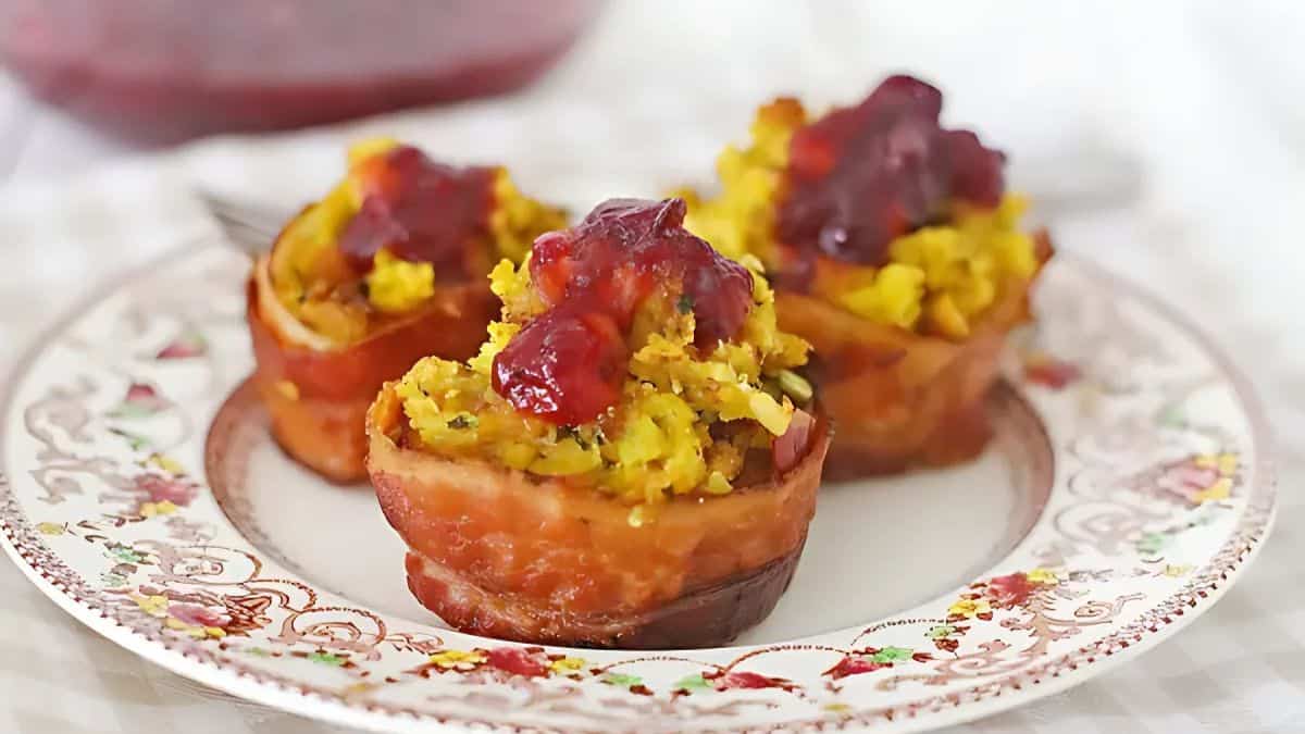 Three bacon-wrapped stuffing muffins topped with cranberry sauce are presented on a decorative plate. The muffins appear golden brown, and a blurred background shows a partial glimpse of additional cranberry sauce.