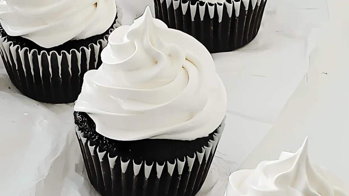Four chocolate cupcakes with swirls of white frosting on top sit on a white surface. The cupcakes are arranged in a slightly scattered manner, highlighting the contrast between the dark chocolate and bright frosting.