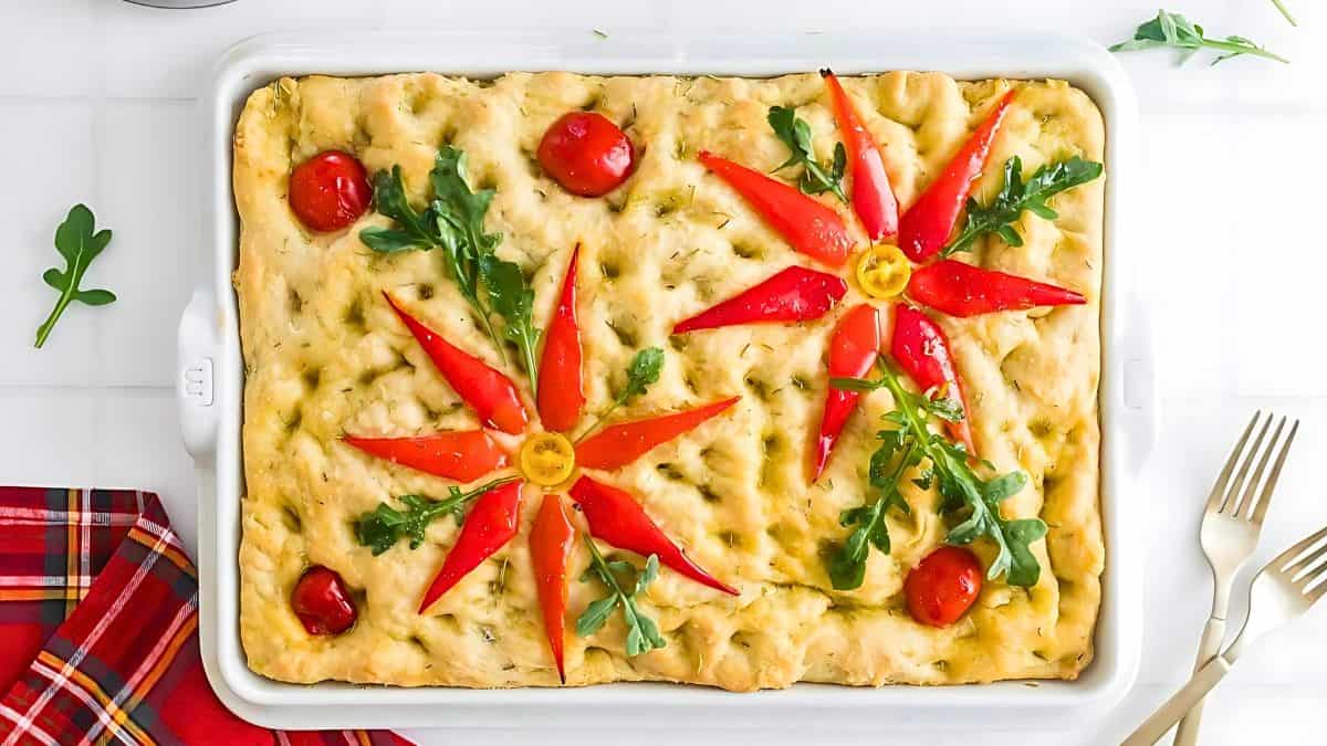 Rectangular focaccia bread topped with red pepper strips, cherry tomatoes, and arugula styled like flowers. The bread is in a white dish, set on a white table. Plaid red napkins and two gold forks are beside it.