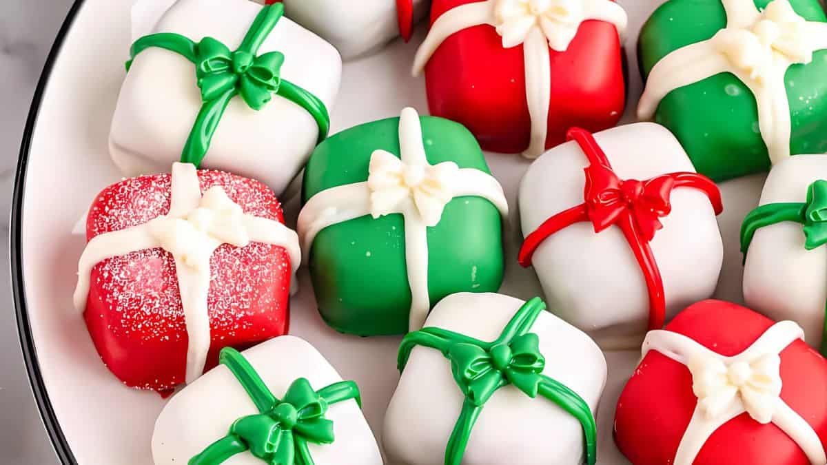 An assortment of small, square cakes decorated as gift boxes with red, green, and white icing. Each cake features a bow on top, and one red box is sprinkled with sugar. They are arranged on a white plate.