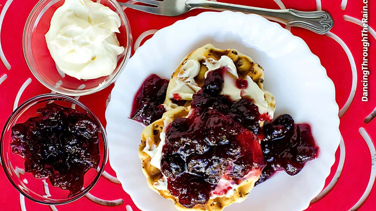 A white plate with waffles topped with whipped cream and cherry compote sits on a red table. A fork is placed nearby. Accompanying small bowls contain more whipped cream and cherry compote.