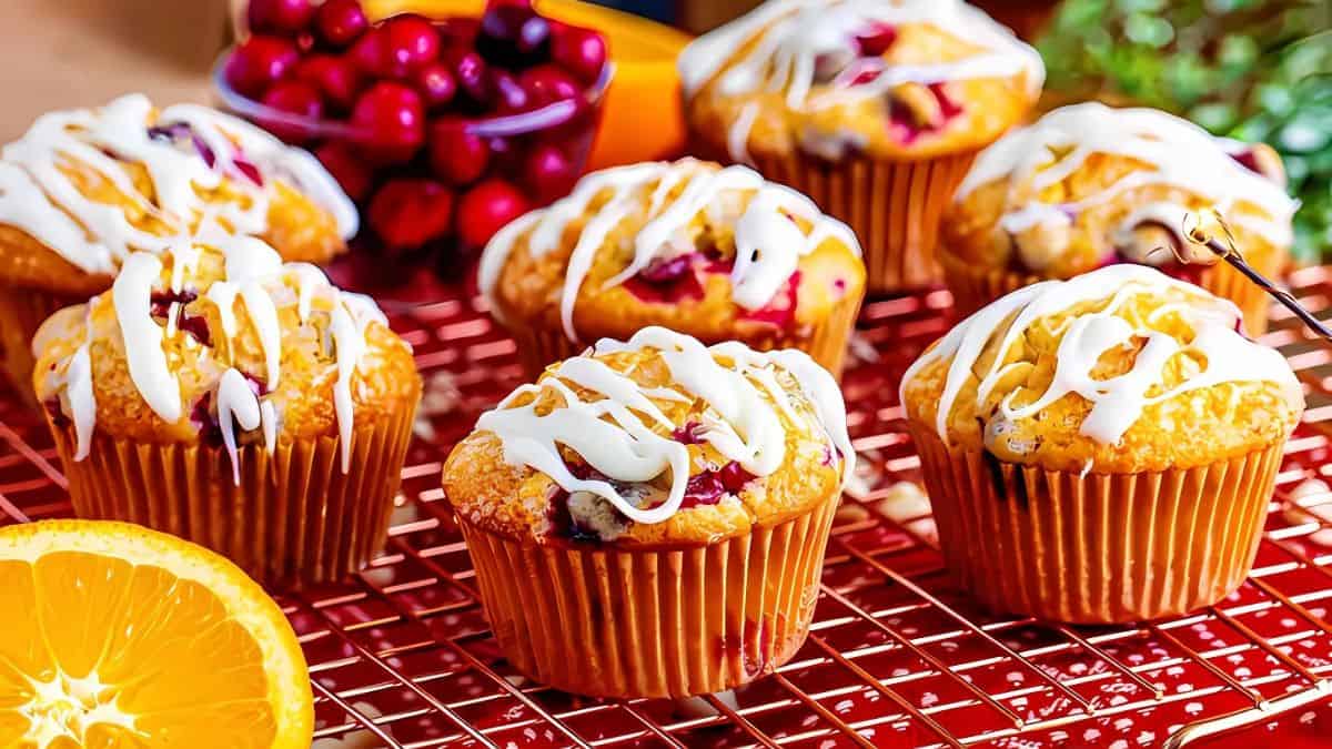 Muffins with cranberries and white icing sit on a wire rack. A sliced orange and a bowl of cranberries are in the background, adding a vibrant touch to the scene.