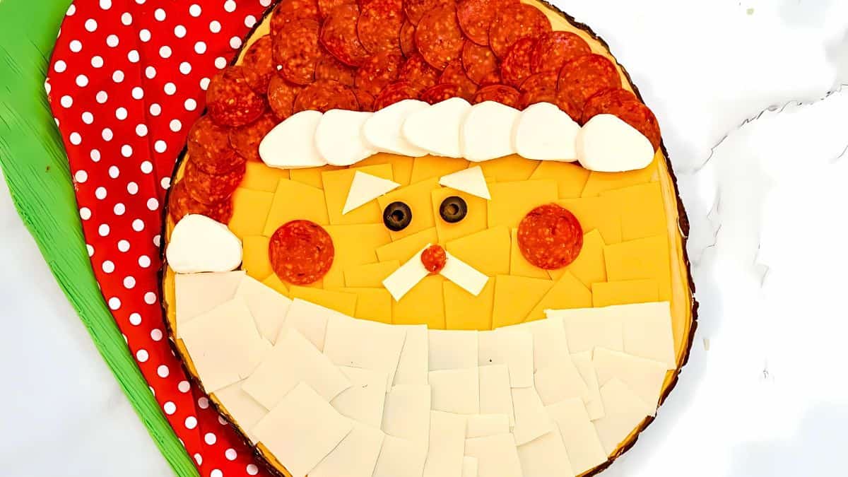 A festive Santa face made of sliced cheese, pepperoni, and mozzarella, arranged on a round platter. The red hat is topped with mozzarella rounds, and the beard is formed with cheese slices. A red and green napkin is on the side.
