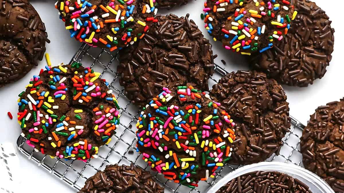Chocolate cookies topped with colorful and chocolate sprinkles are spread across a cooling rack. Some cookies are also placed directly on a white background, with additional chocolate sprinkles nearby.