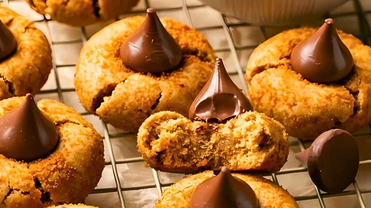 Cookies with chocolate kisses on top are arranged on a cooling rack. One cookie is split in half, showing its soft interior. The cookies are golden brown with a crumbly texture.