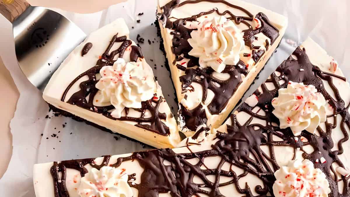 A delicious white chocolate dessert covered in dark chocolate drizzle, topped with whipped cream and crushed peppermint candies. The dessert is sliced and presented on a sheet of parchment paper with a cake server beside it.
