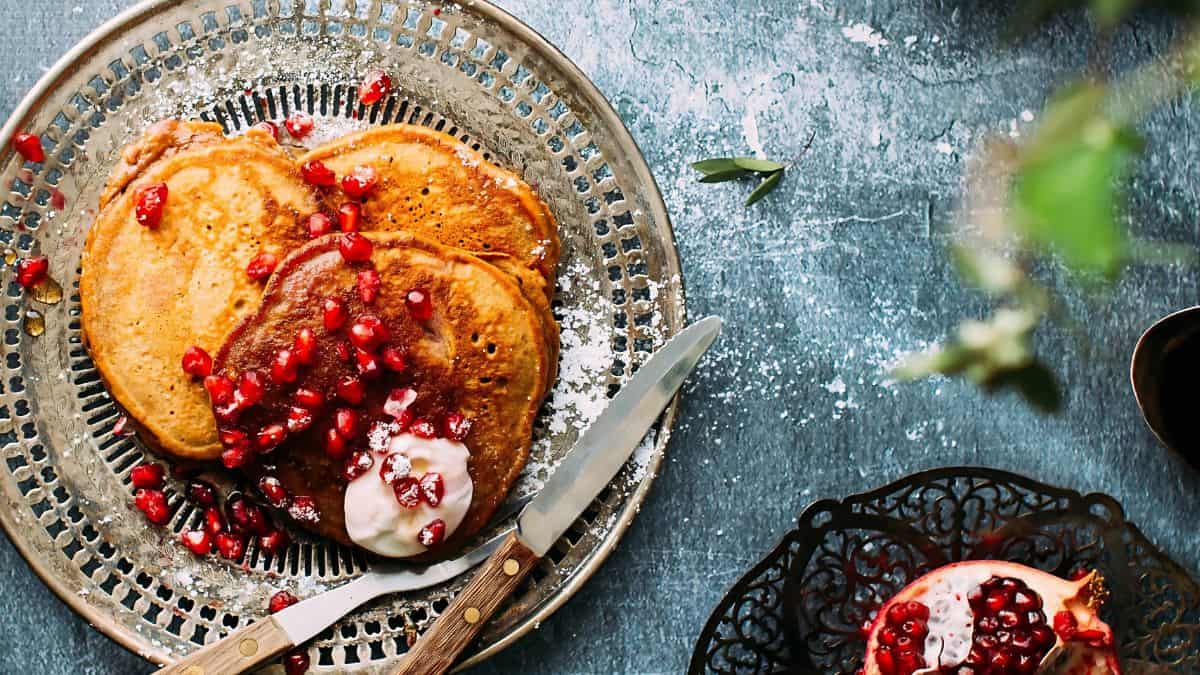 A plate of pancakes topped with pomegranate seeds and a dollop of cream, accompanied by a knife. Nearby, a bowl contains additional pomegranate seeds. The table is rustic, adding to the cozy setting.