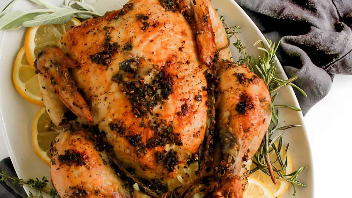 Roasted chicken garnished with herbs and surrounded by lemon slices on a white plate. A dark cloth napkin is partially visible beside the plate.