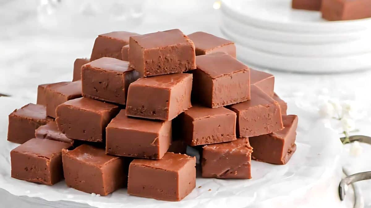 A stack of chocolate fudge squares is arranged on a piece of parchment paper. The fudge appears creamy and smooth, with layers of squares neatly piled on top of each other. Several white plates are visible in the background.
