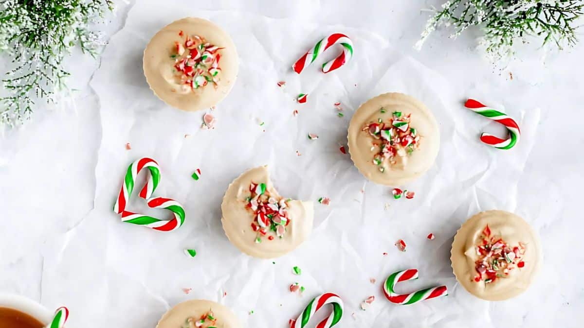Round cookies with white icing and festive red and green sprinkles are scattered on crumpled parchment paper. Small candy canes and sprigs of evergreen add a holiday touch to the scene.