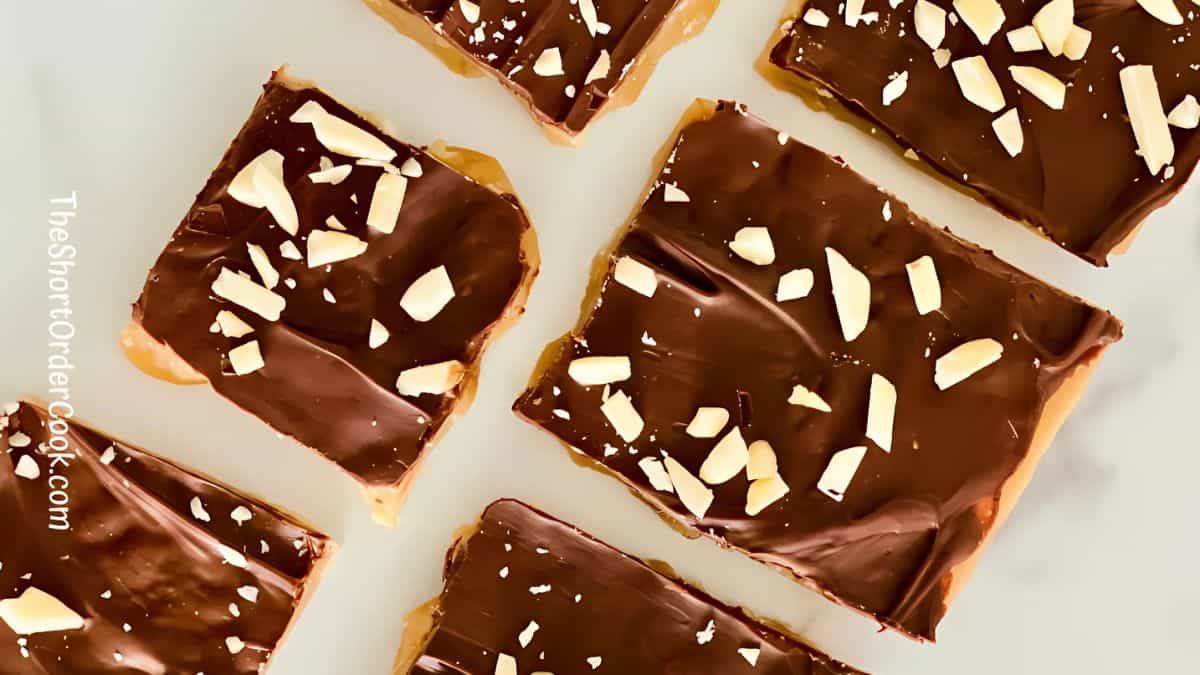 Squares of chocolate toffee topped with chopped nuts are arranged on a white surface. The glossy chocolate contrasts with the crunchy nuts, creating a tempting treat.