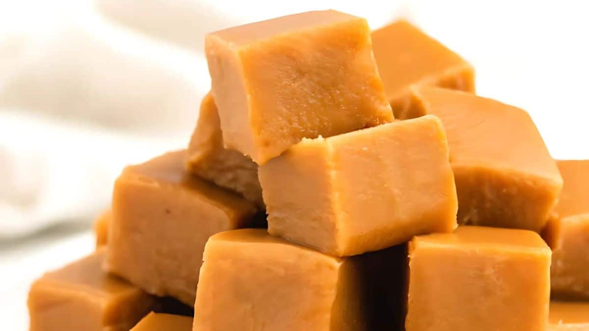A close-up of a stack of creamy caramel fudge cubes, showcasing their smooth and glossy texture. The fudge is arranged neatly, with a soft lighting highlighting its rich, golden-brown color.