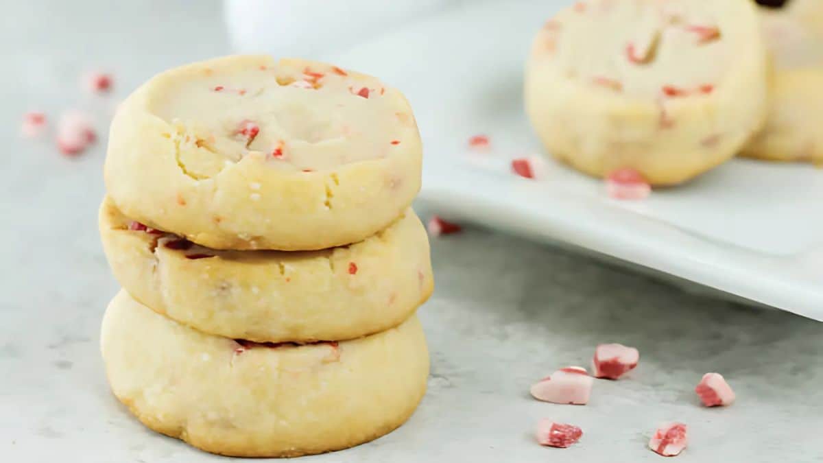 A stack of three shortbread cookies with bits of pink peppermint candy on a light marble surface. More peppermint cookies and candy pieces are scattered in the background on a white plate.
